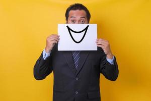 hombre de negocios con ojos anchos cubriendo la mitad de su cara con un emoticono de sonrisa en papel, aislado en fondo amarillo foto