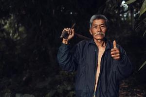 el retrato de un agricultor asiático muestra los pulgares hacia arriba y lleva una azada contra un fondo oscuro vintage. foto