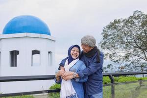 Asian elderly couple in casual style is smiling and looking each other, old man hugging his wife and hold hands each other in front of aesthetic dome and trees. photo