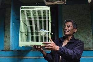 Asian elderly man holding green bird cage outside traditional wooden house. photo