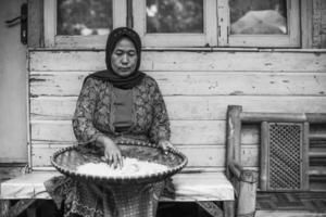 El retrato en blanco y negro de una mujer local asiática está sentado y tamizando arroz con un colador de bambú contra una casa tradicional de madera. foto