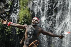 Expressive Papua man wearing traditional clothes of Dani tribe, red-white headband and bangle is holding little Indonesia flag and celebrating Indonesia independence day against waterfall background. photo