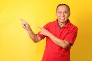 un anciano asiático con una camiseta informal de pie y sonriendo aislado en un fondo amarillo apuntando con la mano y el dedo hacia un lado con una expresión feliz y natural en la cara. foto