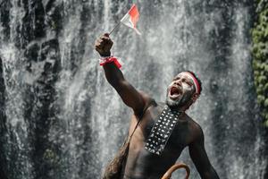 Papua man of Dani tribe say merdeka, celebrating Indonesia independence day against waterfall background. photo