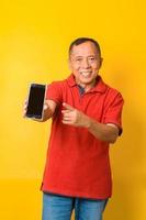 Photo of Asian old man holding an empty blank screen phone with direct finger pointing to screen wearing red t-shirt isolated on yellow color background