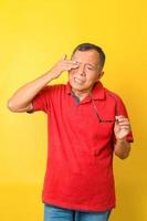 Asian senior man feels tired after reading with eye glasses isolated on yellow background. Old man with eye irritation, optical problem. photo