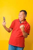 Photo portrait of Asian cheerful old man celebrating victory holding phone in one hand isolated on yellow colored background.