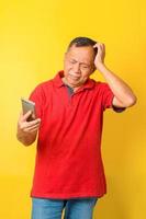 Photo of Asian old man feeling confused and dizzy while looking to smartphone  wearing red t-shirt isolated on yellow color background