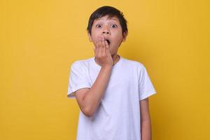 Asian boy in a casual style feeling suprised and closing mouth by hand, on yellow background. Mock up for kids fashion. photo