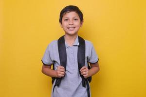 Asian school boy in casual style with black backpack is smiling isolated on yellow background. Back to school concept. photo