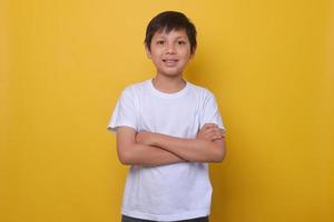 Attractive Asian boy in white casual t-shirt is smiling and folding arms isolated on yellow background. photo