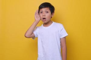 Asian boy with hand directing ear listening hearing words isolated on yellow background. Concept what kids hear. Overhear expression. Mock up for kids fashion. photo