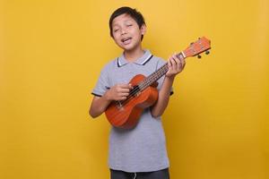 Happy funny asian in grey polo shirt boy sing a song and playing ukulele against yellow background. photo