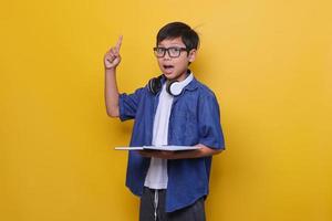 niño inteligente con ropa informal, auriculares y anteojos sosteniendo un libro y señalando con el dedo índice sobre fondo amarillo. aha momentos, teniendo idea. foto