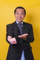 Attractive Asian man in business suit standing against yellow background, presenting and showing money one hundred thousand rupiah. Financial and savings concept. photo