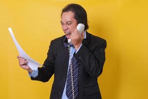 Asian businessman shouting as he holds out phone when see report paper, isolated on yellow background. photo