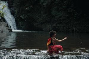 wamena, papua, indonesia, 01 de junio de 2022 atractiva joven papua de la tribu dani vestida con ropa tradicional está sentada y jugando agua contra el fondo de la naturaleza. foto