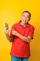 Photo of Asian old man smiling with wide eyes and holding smartphone with direct finger pointing to screen wearing red t-shirt isolated on yellow color background