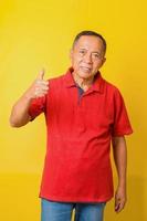 Asian senior man wearing casual t-shirt standing over isolated yellow background doing like thumbs up gesture with hand. Approving expression looking at the camera showing success. photo
