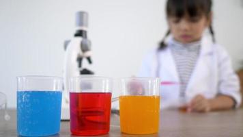 foco seletivo em líquidos de três cores em copos, borrão menina asiática vestindo casaco usando conta-gotas para sugar o líquido do copo de vidro no fundo. enquanto estudava química científica video