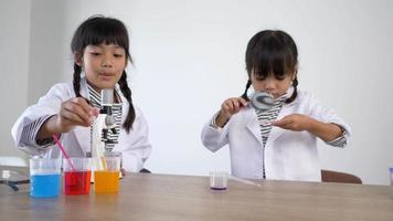 Two Asian siblings wearing coat in lap, girl use dropper to suck liquid from glass beaker and use magnifying glass looking at blue liquid on Petri dish, studying science chemistry with fun video
