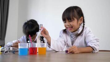 Two Asian siblings wearing coat in lap, girl use dropper to suck liquid from glass beaker and use magnifying glass looking at blue liquid on Petri dish, studying science chemistry with fun video