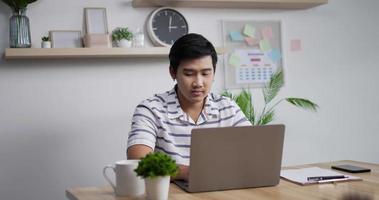 retrato de un hombre de negocios asiático pensativo que trabaja en una computadora portátil en la oficina en casa. joven gerente masculino mirando la pantalla de un portátil en un lugar de trabajo remoto.