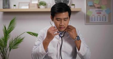 porträtt av leende ung asiatisk manlig läkare kardiolog bär vit medicinsk rock visar stetoskop och tittar på kameran. medicinsk och hälsovård koncept. video