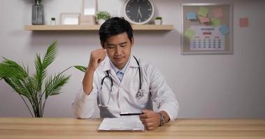Portrait of Tired young asian male doctor cardiologist wearing white medical coat and stethoscope sitting at desk. Medical and health care concept.