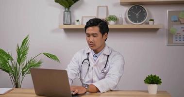 portrait d'un jeune médecin cardiologue asiatique souriant portant un manteau médical blanc tapant un ordinateur portable et montrant le pouce vers le haut dans le bureau de la clinique. concept médical et de soins de santé.