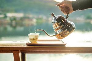 Hand holding vintage teapot and pouring hot tea to cup on wood table against lake view background at coffee shop in the morning sunrise, Ban Rak Thai village, Mae Hong Son province, Thailand photo