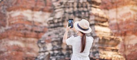 Happy tourist woman wearing surgical face mask and taking Photo by mobile smartphone, protection COVID-19 pandemic during visiting in Ayutthaya temple. new normal, safety travel and travel concept