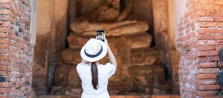 Happy tourist woman in white dress taking Photo by mobile smartphone, during visiting in Wat Chaiwatthanaram temple in Ayutthaya Historical Park, summer, solo, Asia and Thailand travel concept