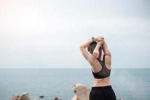 Young fitness female in sportswear stretching body against ocean view, healthy woman exercise in morning. Workout, wellness and work life balance concepts photo