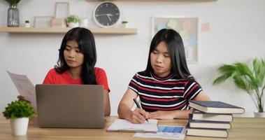 retrato de dos colegialas asiáticas que estudian en línea haciendo notas en un cuaderno con una computadora portátil en la mesa en casa. concepto de aprendizaje y educación a distancia. video