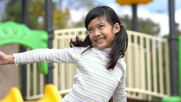 toma en cámara lenta, retrato de una niña asiática de pie cerca del control deslizante en la parte de atrás y juega sola con feliz en el patio de recreo video