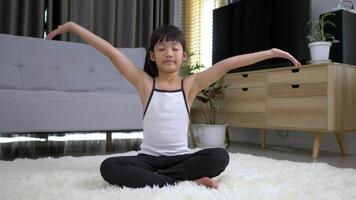 liten asiatisk flicka som sitter på mattan stängde ögat och placerade två händer på bröstet i meditationsasanaställning hemma video