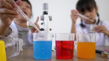 Selective focus at three colors liquids in beakers, blur Asian little girl wearing coat using dropper to suck liquid from glass beaker in background. while studying science chemistry video