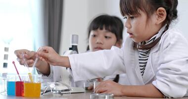 Selective focus at Asian little girl wearing coat using dropper to suck liquid from glass beaker, blur three colors liquids in beakers, while studying science chemistry video