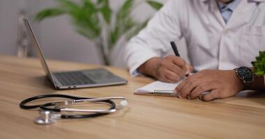 un gros plan d'un stéthoscope est placé sur une table avec un médecin travaillant sur un ordinateur portable et prenant note de l'arrière-plan. notion d'équipement médical video