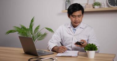 Portrait of young asian male doctor cardiologist wearing white medical coat and stethoscope talking on phone and sitting in clinic. Male has mobile conversation about disease and looks with smile. video