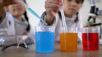 Selective focus at three colors liquids in beakers, blur Asian little girls wearing coat using dropper to suck liquid from glass beaker in background. while studying science chemistry