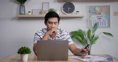 retrato de un hombre de negocios asiático pensativo que trabaja en una computadora portátil en la oficina en casa. joven gerente masculino mirando la pantalla de un portátil en un lugar de trabajo remoto. video