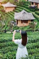 Happy tourist woman in white dress taking photo by mobile smartphone in beautiful Tea garden.Traveler visiting in Ban Rak Thai village, Mae Hong Son, Thailand. travel, vacation and holiday concept