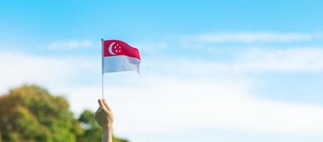 mano sosteniendo la bandera de singapur sobre fondo de cielo azul. día nacional de singapur y conceptos de celebración feliz foto