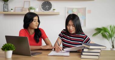 porträtt av två asiatiska skolflickor som studerar online på bärbar dator och gör anteckningar i copybook hemma. distansundervisning och utbildning koncept. video