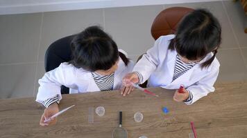 Top view shot, Two Asian siblings wearing coat and clear glasses are using the device for experimenting with liquids. They talking while studying science chemistry with fun video