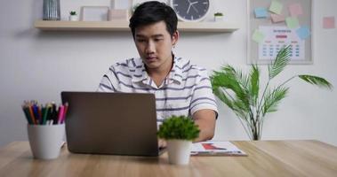 retrato de feliz empresário asiático com computador portátil trabalhando em home office. jovem estudante freelancer masculino usando laptop estudando online trabalhando em casa. video