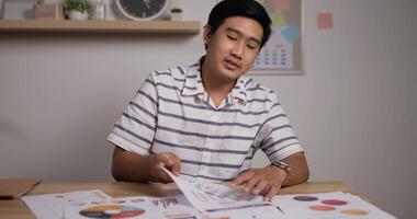 Portrait of Asian bussinesman working through documents on his computer and shows paperwork statistics in online video conference at home office.