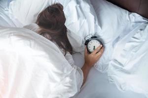 alarm clock and asian woman hand stop time in bed while sleeping, young adult female wake up late in the morning. Relaxing , sleepy, daily routine and have a nice day concepts photo